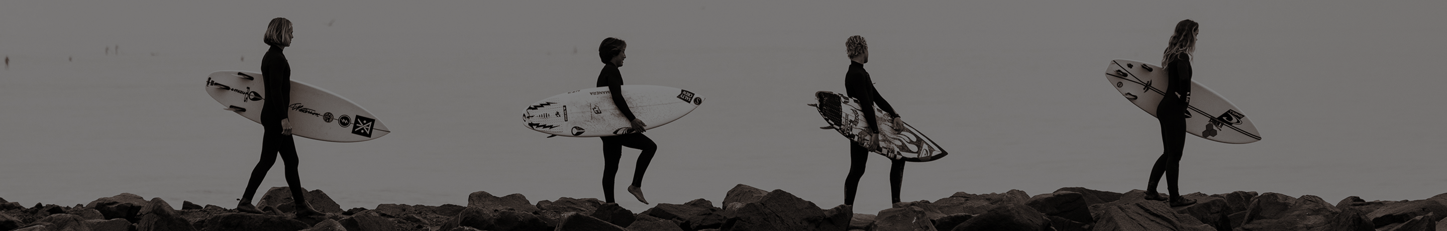 Four surfboarders walk along the ocean while holding their surfboards.